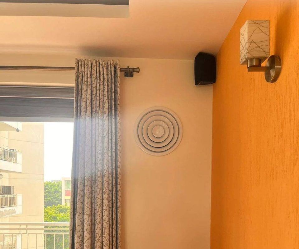 A round diffuser high on an indoor wall, between the curtains and an orange feature wall