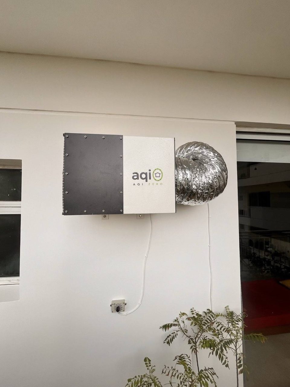 The aqi0 unit, black front plate and white body, wall-mounted on a balcony with its silver flexible duct